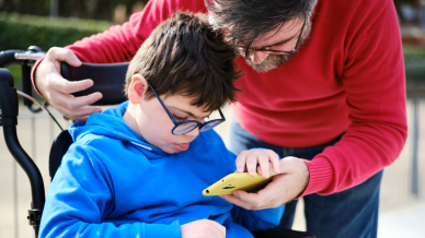Father in a red jumper is with his young wheelchair user son in a blue hoodie. Both looking at a phone held by the father.