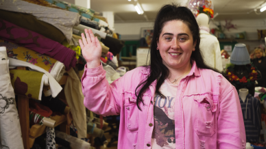 Alex waving and smiling at the camera wearing a pink jacket standing next to rolls of fabric