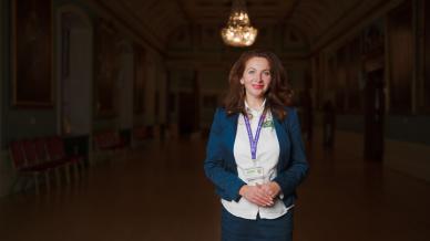 A woman with long curly hair stands in a dimly lit, ornate hall, wearing a blue blazer, white shirt, and lanyard. The hall features chandeliers and portraits on the walls.