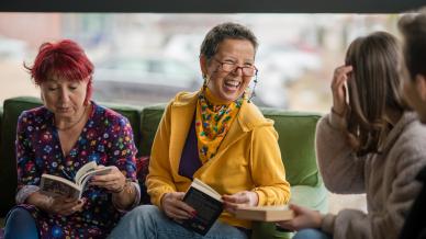 Three women sat on a green couch reading books and conversing